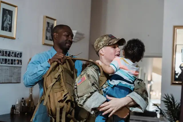 Soldier in uniform holding a young child while a partner helps pack a duffel bag at home