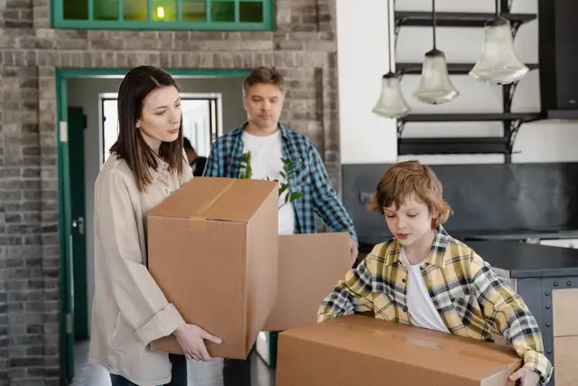 Family of three carrying cardboard moving boxes into the hallway of their new home