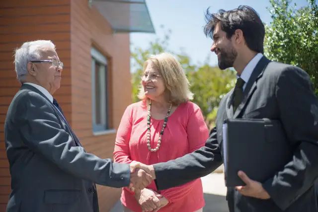 Agent in a dark suit shaking hands with a smiling couple in their sixties outside their home